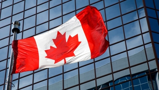 A Canadian flag on a silver pole waving against a dark glass corporate building exterior under blue sky