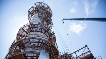 Large industrial column with surrounding scaffolding viewed from below, crane arm extending into a blue sky