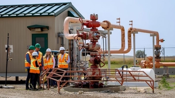 Engineers in orange safety gear and white hard hats inspecting wellhead equipment beside a site building
