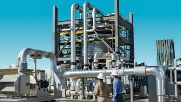Engineers in hard hats examining industrial pipework and equipment outside a large steel processing structure